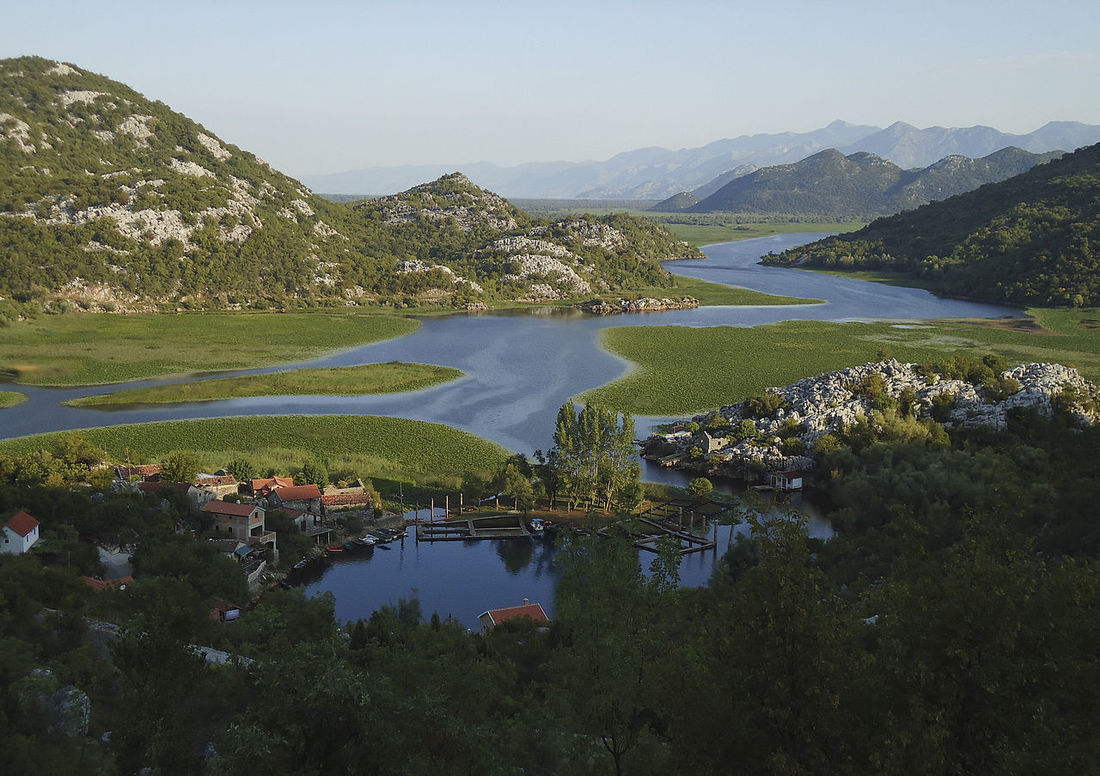 Karuč. Naselje se smjestilo uz estuarij Rijeke Crnojevića, koja utječe u Skadarsko jezero (u pozadini). Na horizontu se uzdiže masiv planine Rumije